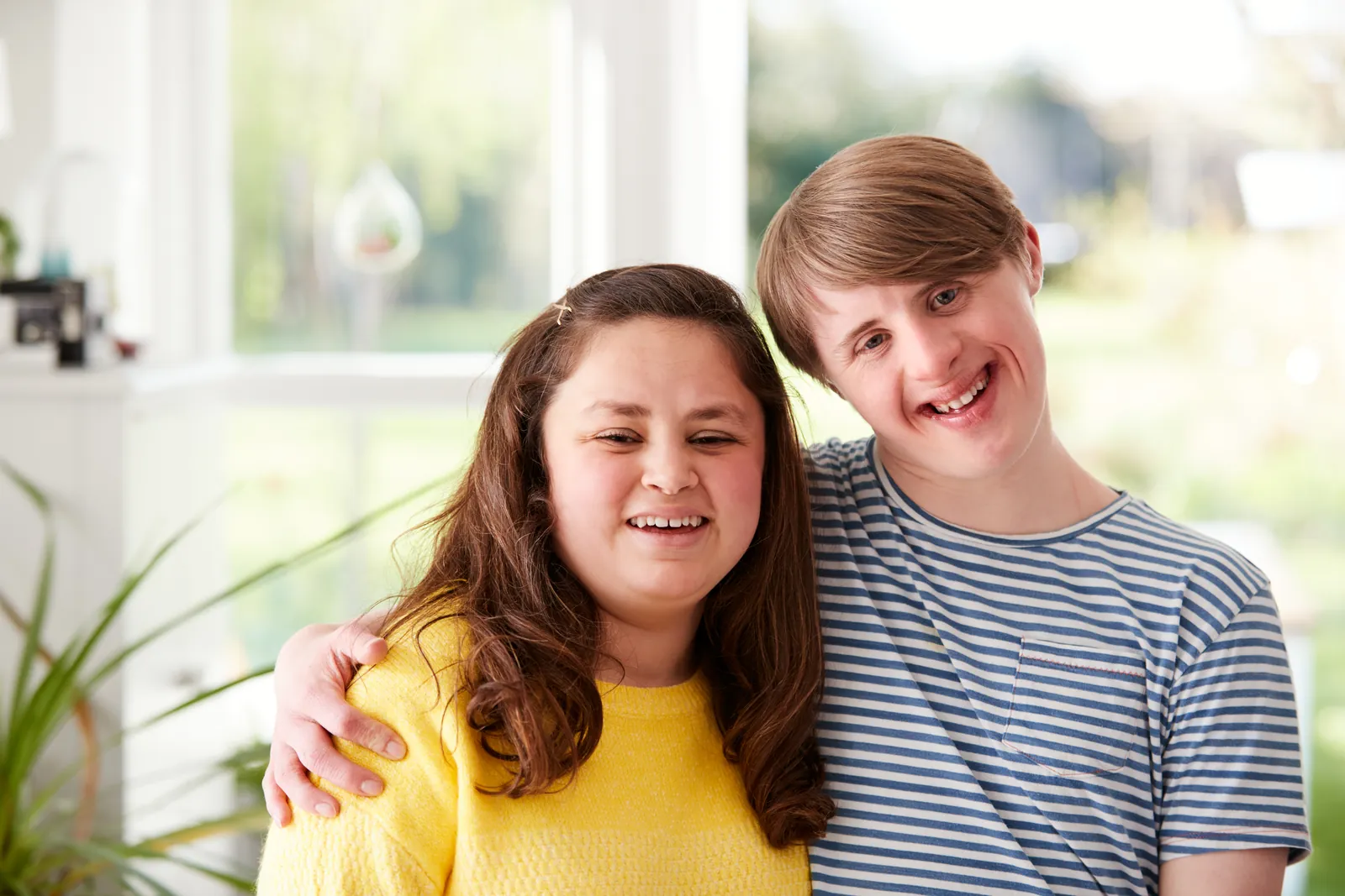 Two smiling friends with Down syndrome standing arm in arm by a sunny window
