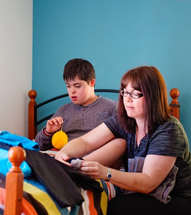 Mother helping her son of 12 years old with Down Syndrome in daily lives to get dressed. He is sit on his bed on the time timers is placed on the side table to stimulate it. He is looking away. Photo was taken in Quebec Canada.